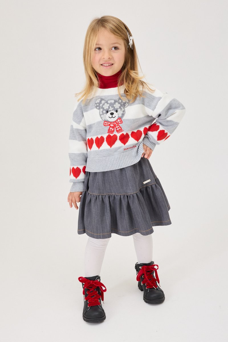 Jeune fille portant un pull rayé avec un ours en peluche et des cœurs rouges, une jupe grise, des collants blancs et des bottes noires avec des lacets rouges, debout la main sur la hanche.