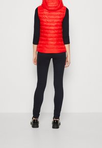 Červená nadýchaná vesta s kapucí, prošívaná textura, kombinovaná s černými úzkými legínami a černými botami. Jednoduchý design, bez viditelných vzorů nebo ozdob.