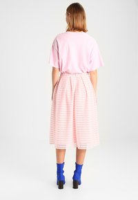 Mujer con cabello hasta los hombros, vestida con una blusa de manga corta color rosa claro, falda midi plisada rosa y botas bajas azules, de pie mirando hacia atrás.