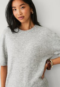 Lys grå sweater lavet af blødt strikket stof med rund hals og korte ærmer. Bemærkelsesværdige guld- og brune armbånd om håndleddet.