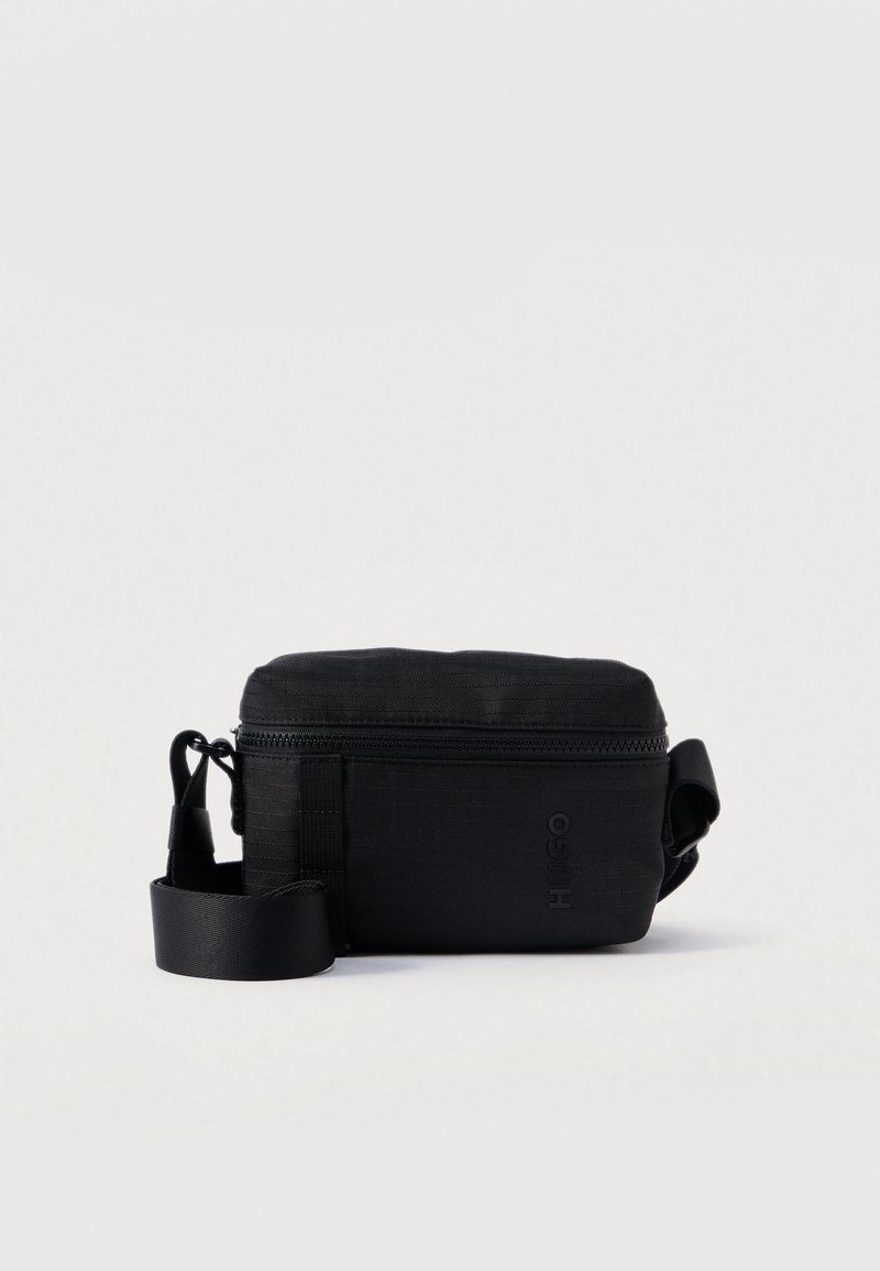 Συμπαγής μαύρη τσάντα χιαστί με κλείσιμο φερμουάρ, ρυθμιζόμενος ιμάντας και το λογότυπο "HUGO" τυπωμένο μπροστά σε απλό φόντο.