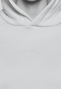 Ljust grå hoodietröja med känguruficka och ett broderat hängarsymbol på bröstet. Tillverkad av mjukt, texturerat tyg.