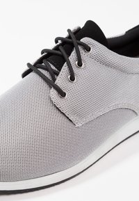 Zapato casual de tela de malla gris con cordones negros, lengüeta negra, ojales plateados y suela blanca con borde negro, mostrado en un ángulo de primer plano.