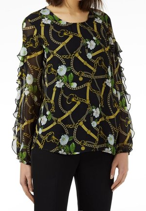 Blusa negra transparente con cadena dorada y estampado floral blanco, con mangas largas con volantes, combinada con pantalones negros.