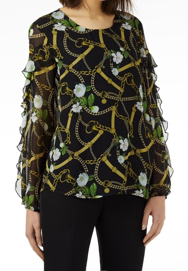 Blouse noire transparente avec chaîne dorée et imprimé floral blanc, manches longues volantées, portée avec un pantalon noir.
