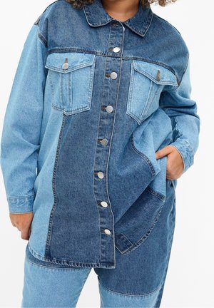 Personne portant une veste en denim bicolore oversize et un pantalon assorti, main dans la poche de la veste, debout devant un fond blanc.