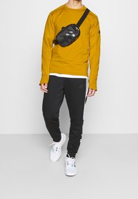 Camicia a maniche lunghe color senape, pantaloni neri con strisce bianche, sneakers bianche e una borsa a tracolla nera con accenti riflettenti.