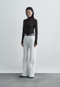 Pulover maro cu guler înalt, pantaloni albi cu croială largă și buzunare frontale, curea maro, pantofi cu toc negri cu vârf ascuțit, fundal minimalist.