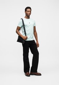 Ανοιχτό μπλε βαμβακερό μπλουζάκι με λογότυπο, μαύρο παντελόνι, καφέ μποτάκια μέχρι τον αστράγαλο και μαύρη τσάντα ώμου, στέκεται μπροστά σε ανοιχτό φόντο.