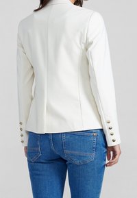 Vit kavaj med en figursydd design, med en söm på ryggen, guldknappstillbehör på ärmarna, kombinerat med blå denimjeans.