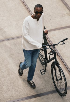 Homme portant des lunettes de soleil, une chemise blanche à manches longues et un jean marchant en tenant un vélo noir sur une surface pavée.