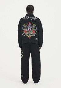 Giubbotto di denim nero con grafiche ricamate colorate sul retro, inclusi un'aquila e teschi, con testo bianco e vestibilità casual.