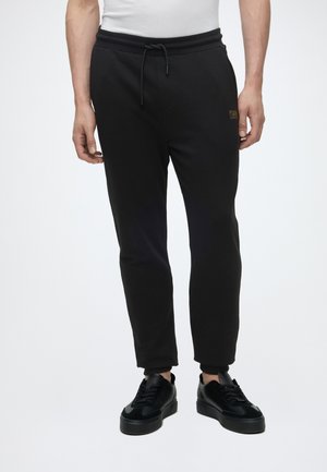Mann trägt schwarze enganliegende Jogginghosen mit Kordelzug in der Taille und schwarze Sneakers, steht vor einem schlichten weißen Hintergrund.