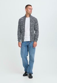 Hombre de pie vistiendo un suéter gris con cremallera, camiseta blanca, jeans azules y zapatillas negras, mirando de lado contra un fondo liso.