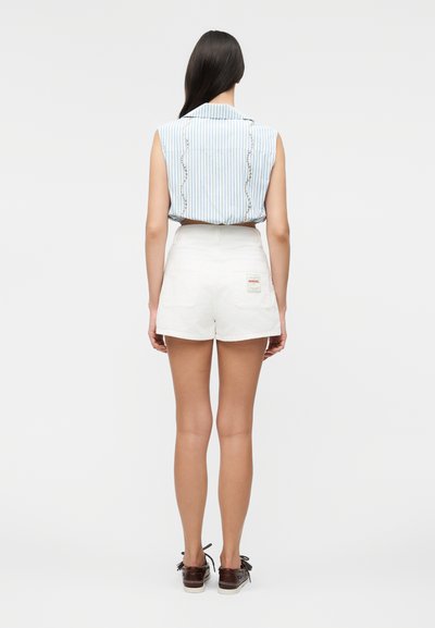 Γυναίκα με μακριά σκούρα μαλλιά, φορώντας μπλε ριγέ αμάνικη μπλούζα και λευκό ψηλόμεσο σορτς με ταμπελάκι στην πίσω τσέπη, στέκεται με την πλάτη.
