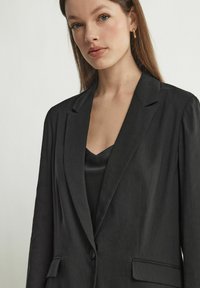 Svart kavaj i lättviktig tyg med skräddarsydd passform, sneddade slag, enkel knappstängning och sidofickor, lager på en svart linne.