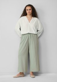 Blouse blanche à manches longues avec un col en V et une coupe décontractée, associée à un pantalon large vert clair avec une ceinture élastique. Ballerines beiges.