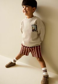 Šviesiai smėlio spalvos džemperis su grafiniu atspaudu, derinamas su raudonai baltų dryžių šortais. Ruda sportinė avalynė ir dryžuotos kojinės užbaigia aprangą.