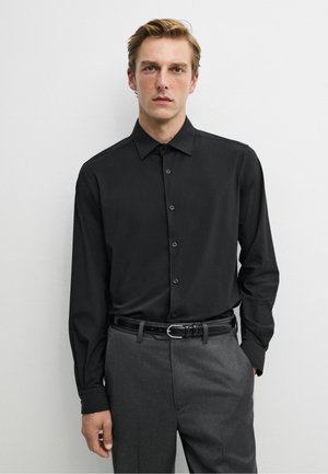 Mann trägt ein schwarzes Hemd mit Knöpfen, in graue Hose mit schwarzem Gürtel gesteckt, steht vor weißem Hintergrund.