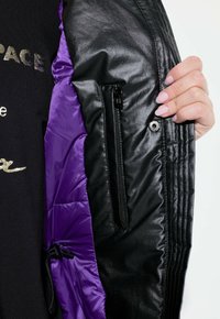 Schwarze Daunenjacke mit lila Innenfutter, Reißverschluss-Tasche und geripptem Textur an denÄrmeln. Das Material wirkt glänzend und isoliert.