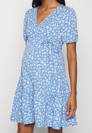 Schwangere Frau in einem blauen Wickelkleid mit weißem Blumenmuster und kurzen Puffärmeln, die mit einer Hand entspannt an ihrer Seite steht.