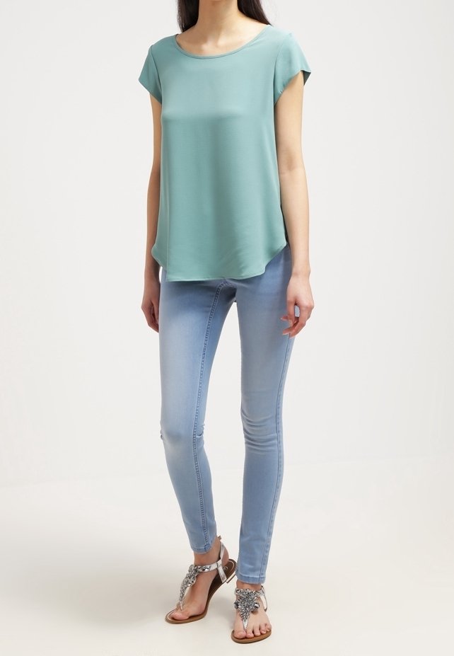 Femme portant un haut ample à manches courtes bleu sarcelle, un jean skinny bleu clair et des sandales argentées ornées de bijoux, à bouts ouverts, debout devant un fond uni.
