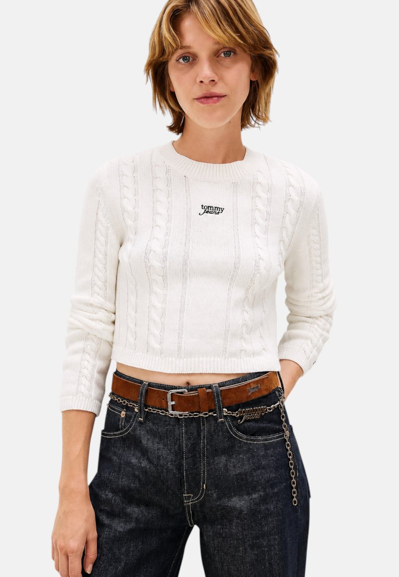 Jeune femme portant un pull court en maille torsadée blanche et un jean foncé avec une ceinture en cuir marron et un accessoire chaîne.