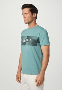 Muž v tyrkysovém tričku s krátkým rukávem, na kterém je černý pruhovaný obdélník a text "HACKETT SPORT", v kombinaci s krémovými kalhotami, stojící čelem vlevo.