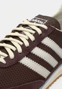 Primo piano di una sneaker maroon Adidas con lacci bianchi e le tre strisce bianche caratteristiche sul pannello laterale in tessuto a pois.