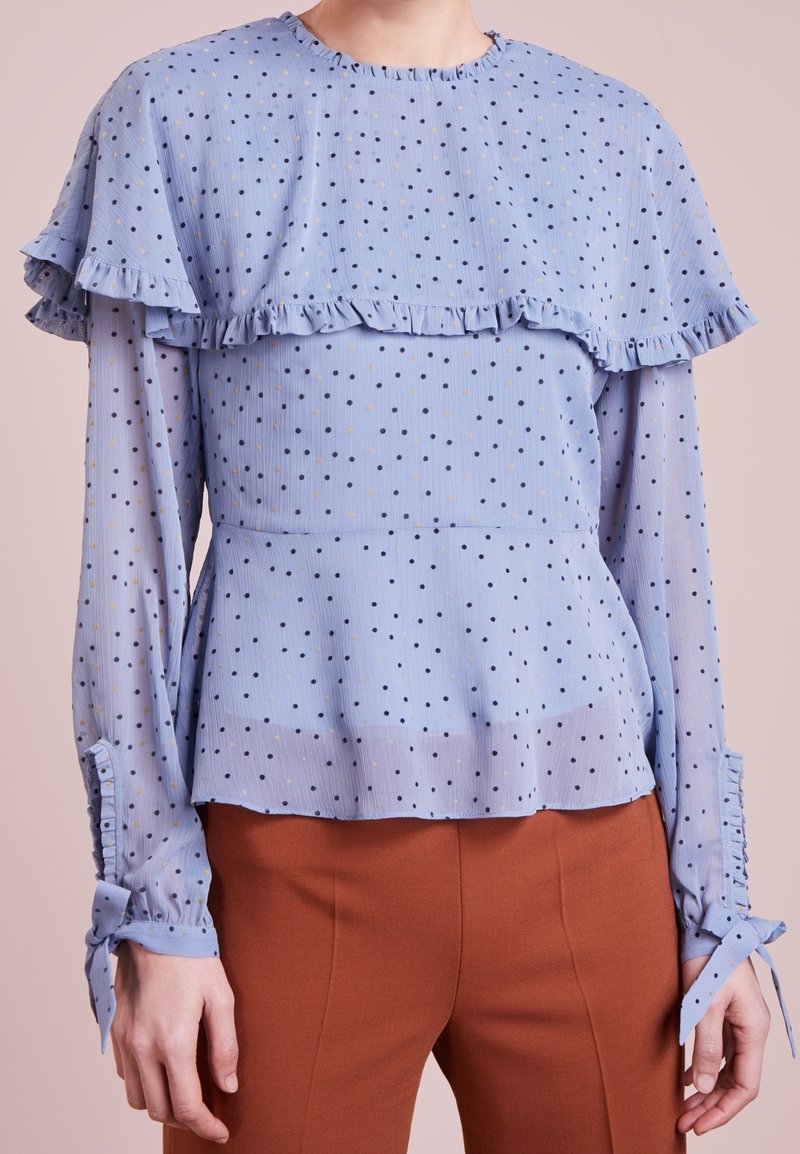Blouse bleu clair avec détail volanté sur les épaules, motif à pois en bleu marine et jaune, manches longues avec poignets noués, tissu texturé.