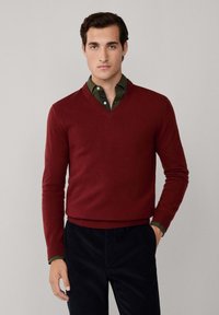 Maglione rosso in lana con scollatura a V, maniche lunghe e polsini a costine. Indossato sopra una camicia con colletto verde scuro. Abbinato a pantaloni scuri.