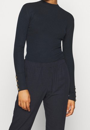 Personne portant un haut noir côtelé cintré à manches longues avec des boutons dorés aux poignets et un pantalon noir ample sur un fond uni.