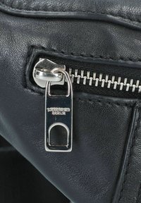 Detail einer schwarzen Lederhandtasche mit einem silbernen Reißverschlussanhänger mit Branding und strukturierter Oberfläche, die Handwerkskunst und Design hervorhebt.