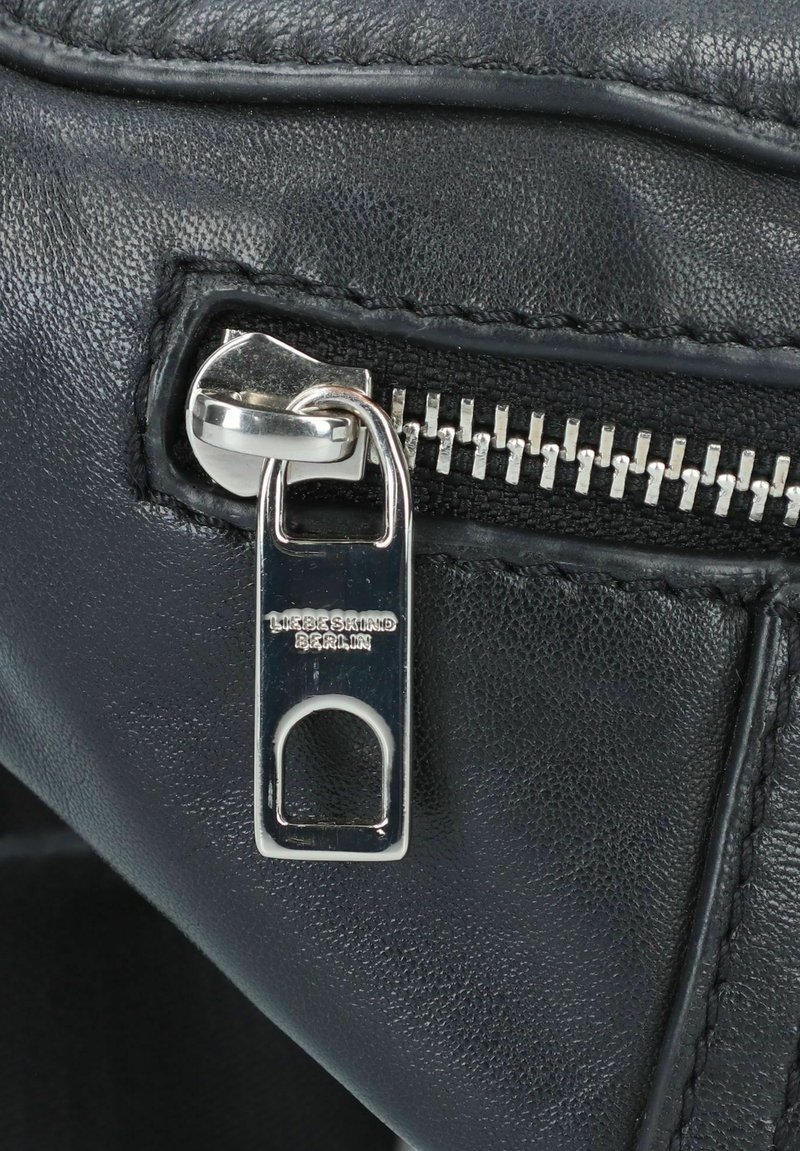 Detail einer schwarzen Lederhandtasche mit einem silbernen Reißverschlussanhänger mit Branding und strukturierter Oberfläche, die Handwerkskunst und Design hervorhebt.