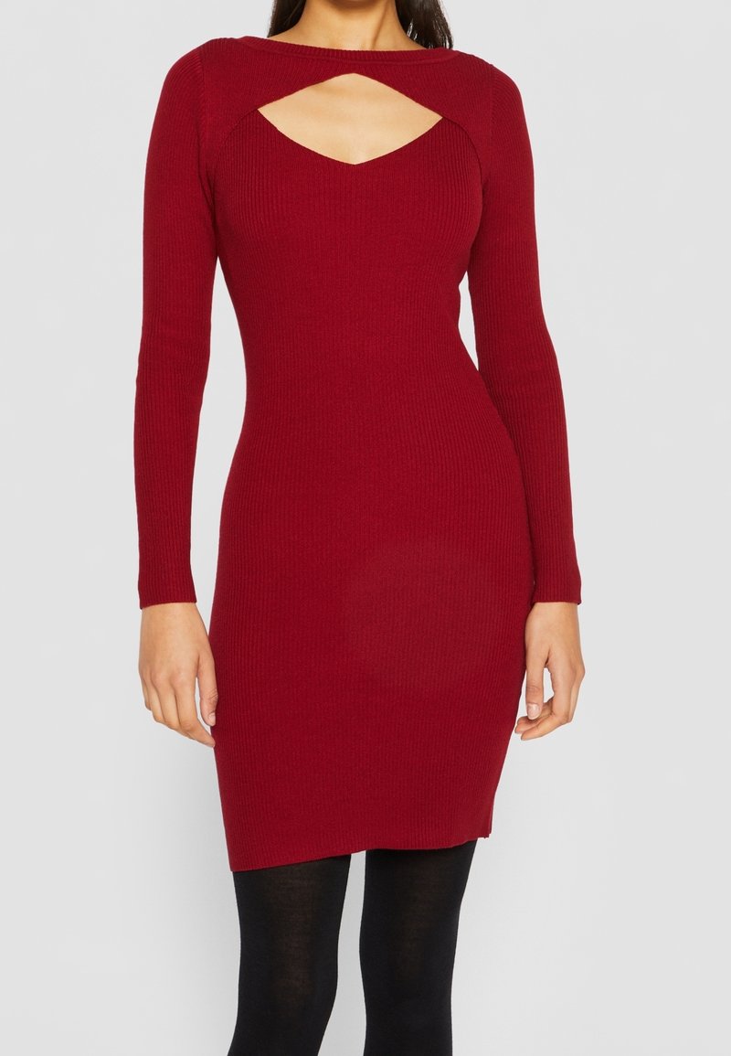 Robe en tricot côtelé de couleur rouge profond avec des manches longues et un décolleté en V plongeant. Design ajusté, arrivant à mi-cuisse. Portée avec des collants noirs.