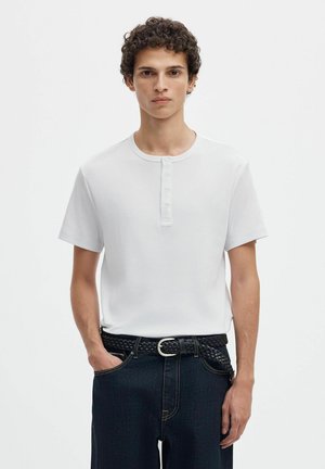 Jeune homme aux cheveux bouclés portant un t-shirt Henley blanc à manches courtes et un jean foncé avec une ceinture tissée noire.