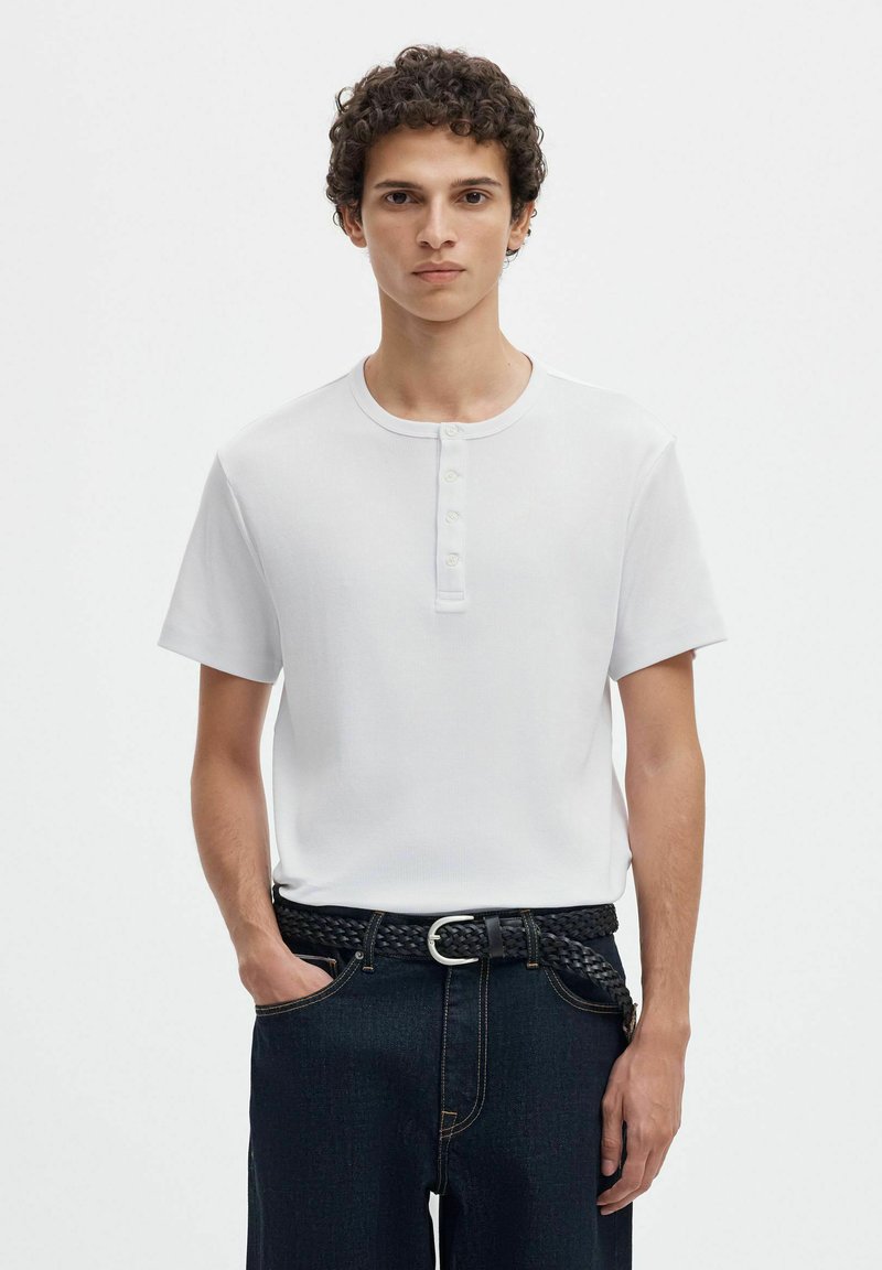 Jeune homme aux cheveux bouclés portant un t-shirt Henley blanc à manches courtes et un jean foncé avec une ceinture tissée noire.