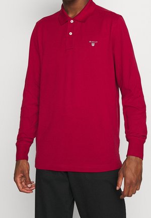 Polo rouge à manches longues avec col et trois boutons, présentant un logo brodé sur la poitrine gauche et une texture coton lisse.