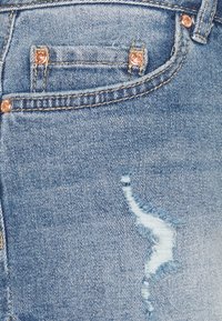 Jeans in denim blu con un design strappato, dotati di rivetti in tonalità dorata e una piccola tasca frontale. La texture è morbida con dettagli sfrangiati.