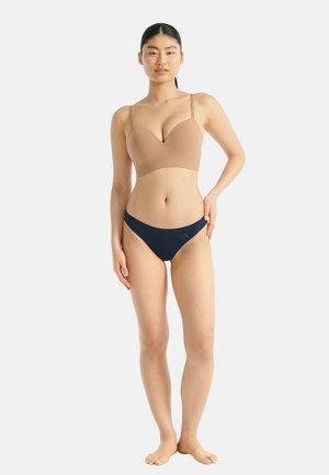 Eine Frau steht barfuß und trägt einen nuden BH und navyblaue Bikinihöschen vor einem schlichten weißen Hintergrund.