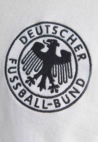Μαύρο κεντητό έμβλημα αετού με το κείμενο "Deutscher Fussball-Bund" σε λευκό υφασμάτινο φόντο.
