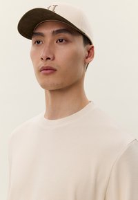 Jauns vīrietis, valkājot bēša krāsas beisbola cepuri un gaišu, teksturētu džemperi ar apaļu kakla izgriezumu, skatās nedaudz uz augšu, fonā ir vienkāršs fons.