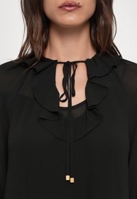 Blusa nera con collettino arricciato con lacci e dettagli dorati. Realizzata in tessuto leggero, ha una consistenza morbida e fluida.