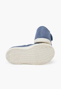 Scarpa di tela blu con punta arrotondata, suola in gomma bianca, grip testurizzato e chiusura con strap adesivo. La vista posteriore mostra i dettagli della cucitura.