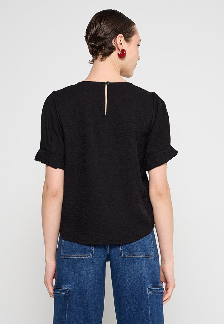 Schwarzes Top aus strukturiertem Stoff, kurze Rüschärmeln, Schlüssellochöffnung auf dem Rücken, getragen mit blauen weit geschnittenen Hosen.