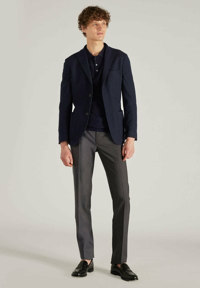 Marineblazer mit Reverskragen, Ein-Knopf-Verschluss und Taschen. Getragen über einem marineblauen Strickpolo und kombiniert mit grauen Hosen und schwarzen Slippern.