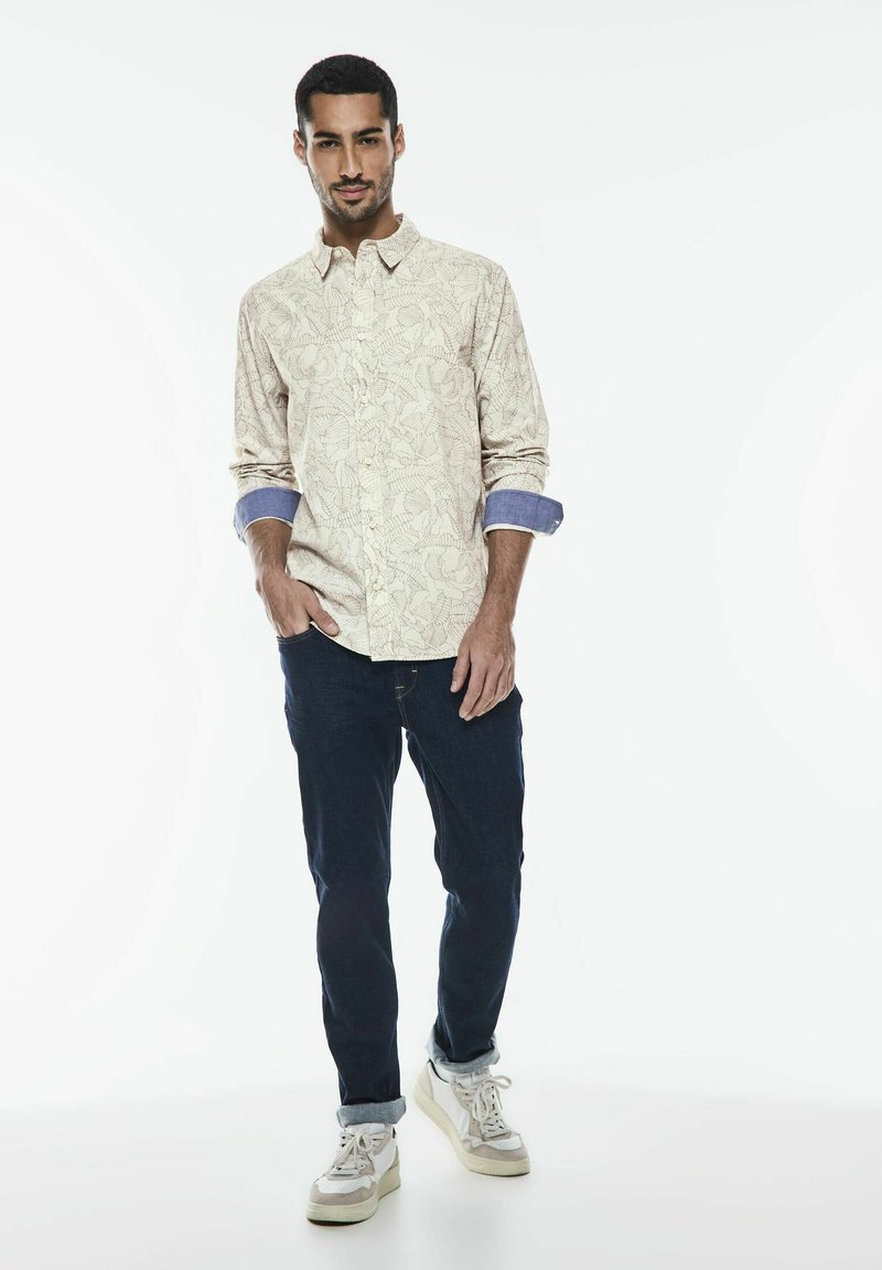 Ljusbeige skjorta med knapp, mönstrad med blommor och denimärmar, kombinerad med mörkblå jeans och vita sneakers med beige detaljer.