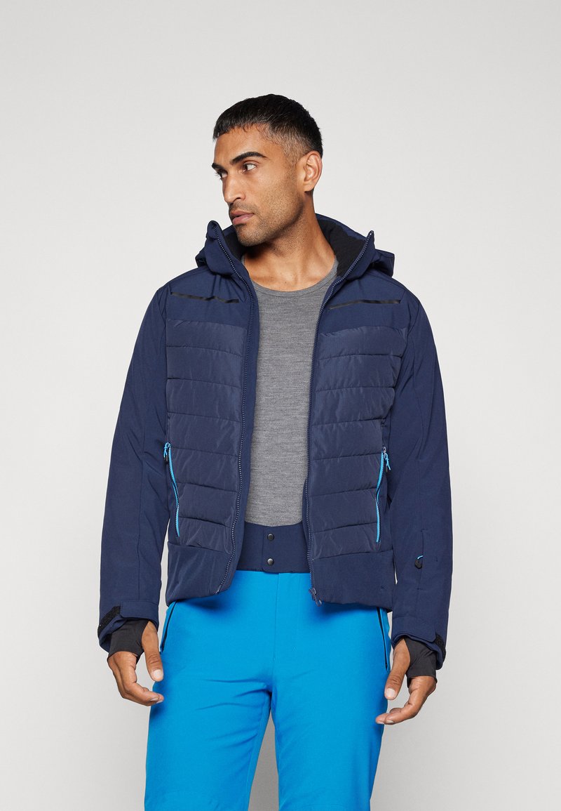 Marine blauwe gewatteerde jas met een ritssluiting aan de voorkant, verstelbare capuchon, ritszakken en contrasterende blauwe accenten. Draagt over een grijs shirt en blauwe broek.