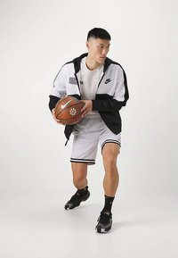 Nike träningsoutfit med en svart och vit jacka med dragkedja, vita shorts med svarta detaljer och svarta sneakers. Håller i en basketboll.