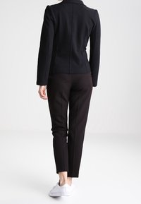 Zwarte slim-fit jas met een hoge kraag, gecombineerd met een slim zwarte broek en witte sneakers, gezien vanuit de achterkant tegen een effen achtergrond.